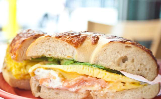Toasters Coffee cup with a bagel that has egg, ham, and avocado on it