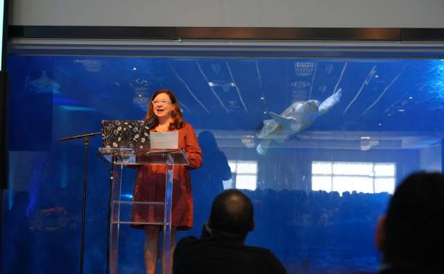 Woman speaking at podium there is an aquarium with a giant sea turtle behind her