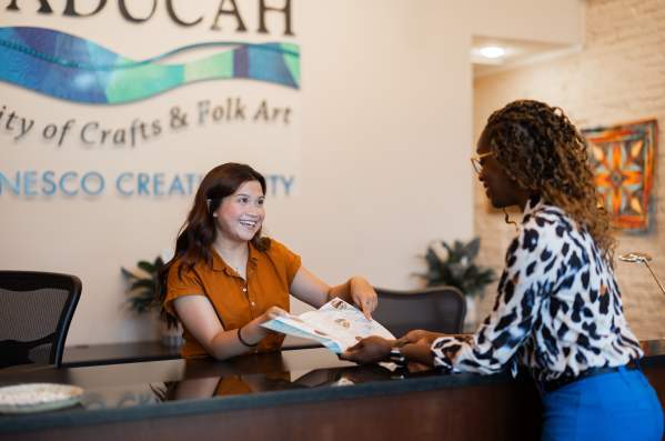 A woman stands behind the counter at the Paducah Convention & Visitors Bureau and helps another woman who is visiting Paducah. They are holding a map and pointing at a location.