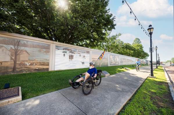 A man pedals a recumbent bike past Paducah's Wall to Wall Floodwall murals. A woman is in the background on a bicycle. The sun shines through the trees.