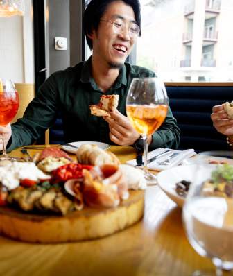 An Asian man holds a slice of pizza while laughing with a woman. The table is surrounded by fresh Italian dishes