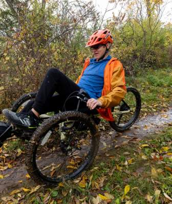 A person in an orange jacket and helmet rides a recumbent mountain bike on a muddy trail surrounded by fall foliage in Middleton, WI, conveying adventure and focus.