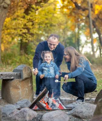 A white man, white woman and young white girl smile while roasting marshmallows over the fire surrounded by fall foliage at Picnic Point