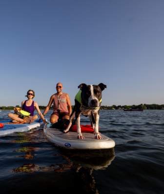 A dog stands on a paddleboard on Lake Monona while two adults ride on paddleboards behind the dog.