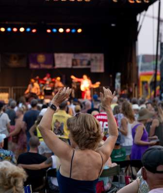 A white woman raisers her hands in as she dances in a crowd in front of a stage where music is being performed at La Fete de Marquette
