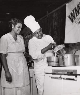 A black and white photo of a Black man, Carson Gulley, cooking on TV with a Black woman.