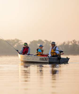 A canoe with three people paddles on calm water during sunset, surrounded by trees and a serene landscape. The scene evokes outdoor adventure.