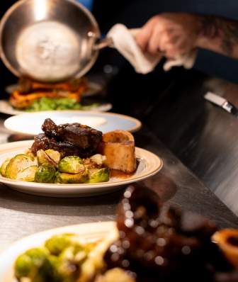 A chef slides food across the counter for a server to take and give to guests. the plate of food has a steak and Brussels sprouts covered in sauce.