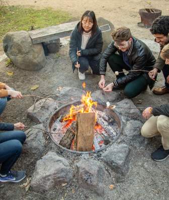 A group of college students gather around a campfire to roast marshmallows