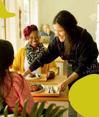 A woman sets a tray of food down on a table between two women
