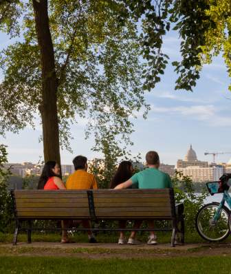 A shot of the city skyline over the lake while a group of people sit on a bench and two others stand next to them with bikes overlooking the views.