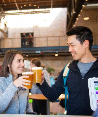 A white woman and Asian man clink beer glasses at a bar inside the Garver Lounge at Garver Feed Mill