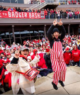 Three women dressed in UW badger gear jumping and cheering with a crowd of Badger fans behind them.