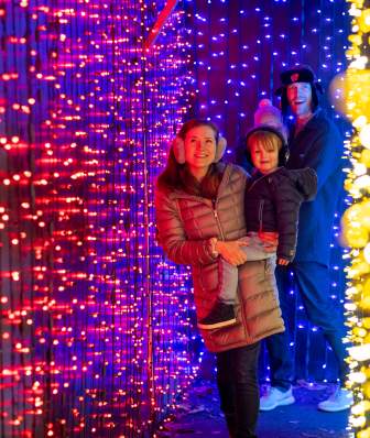 A mom and dad each holding one of their kids as they walk through a tunnel of holiday lights at Henry Vilas Zoo.