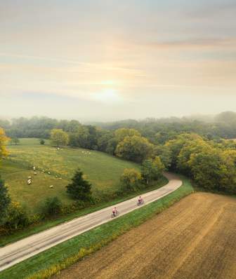 A zoomed out shot of two people bicycling in a rural portion of Madison. The sun is rising over farmland and think tree lines are seen along the horizon
