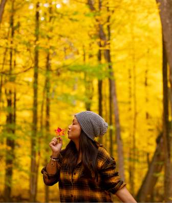 A woman in a plaid shirt and beanie holds a red leaf in a vibrant yellow forest, surrounded by tall trees. The scene captures autumn's beauty.
