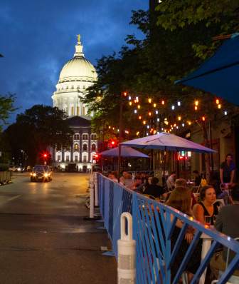 A lively street scene at night, featuring outdoor dining under string lights, with a prominent dome building in the background.