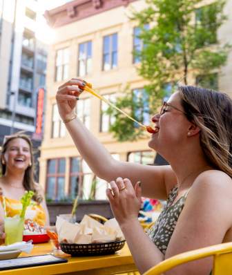 A person sits at an outdoor restaurant patio, holding up a stretchy cheese curd toward their mouth at a yellow table with baskets of food, while other diners and city buildings appear in the background.