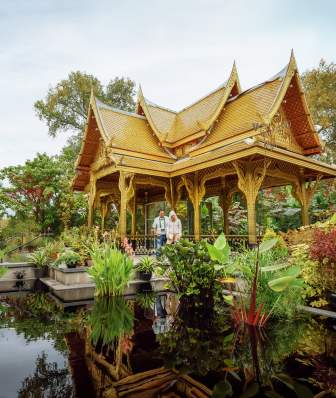 A wide shot of two people standing under the Thai Pavilion at Olbrich Botanical Gardens surrounded by a pond and lush greenery.