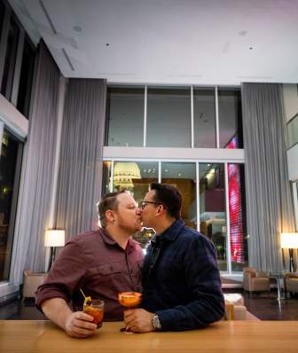 Two men at a hotel bar kiss with the Wisconsin State Capitol building lit up in the background.