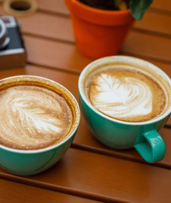 Two mugs with latte art on top, sitting on a wooden table, with a camera in the background.