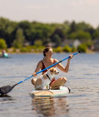A white woman and a medium sized white and brown dog sitting on a paddle board in the lake with rows of trees behind them.