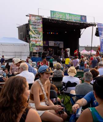 A large crowd sits and stands on a grassy area facing an outdoor concert stage at La Fête de Marquette, with musicians performing under stage lights and festival banners visible above the stage.