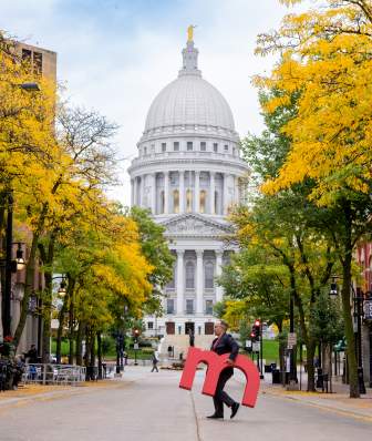 An image of a man walking across State Street in downtown Madison holding a large red "m" with the Capitol building in the background.