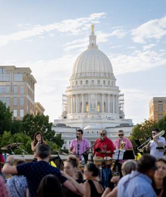 Live band performing before a crowd in an outdoor plaza, with the Wisconsin State Capitol dome rising prominently in the background under a bright sky.