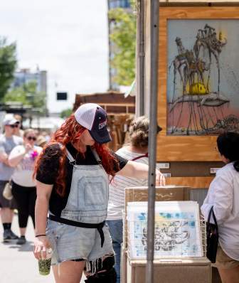 People browsing artwork at an outdoor art fair on a sunny day, with framed surreal paintings displayed in wooden booths and visitors walking past along a city street.