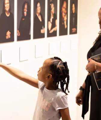 A young Black girl points at the portrait of a Black woman in an art gallery while an adult Black woman stands behind her