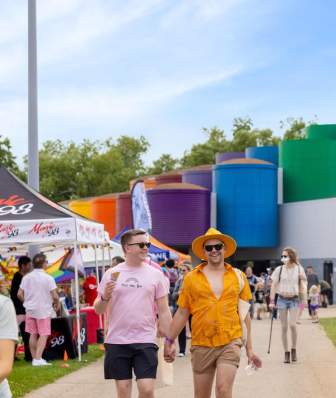 A vibrant outdoor scene with colorful buildings in the background. People stroll along a path, enjoying a lively event with tents and activities.