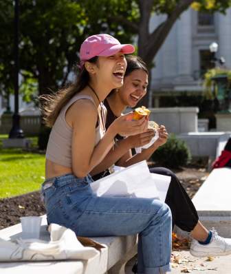 Two Asian women laugh while eating snacks on a bench outside of the Capitol building