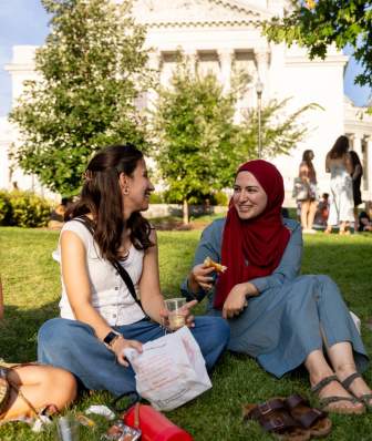 Two women, one wearing a hijab, sit and have a picnic on the lawn in front of the Capitol building