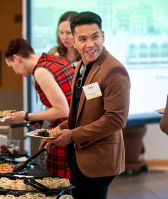 A man smiles at a person out of frame as he adds food to his plate at a buffet line at a meeting.