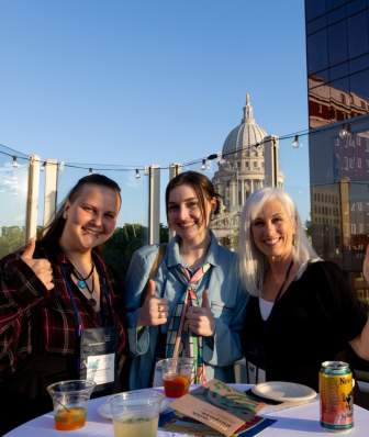 Three white women conference attendees share drinks and thumbs of the camera on the rooftop of the Madison Children's Museum with the State Capitol building in the background