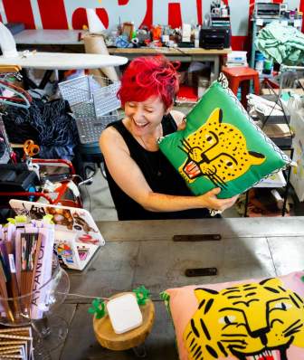 A woman with bright red hair hold sup a brightly colored pillow with a jaguar on it while shopping at Zip Dang