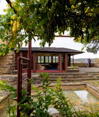 The outdoor courtyard at Frank Lloyd Wright's Taliesin with a reflection pool, Buddhist statue and trees surrounding a room of the home that has large glass windows.