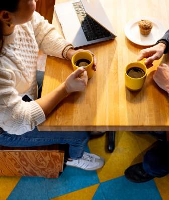 A photo from above of a woman's and a man, both holding coffee, at a table in a coffee shop next to a laptop