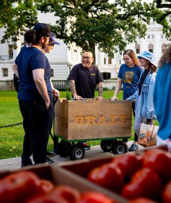 An Asian man, Chef Tory Miller, stands and talks to people around his cart at the Dane County Farmer's Market