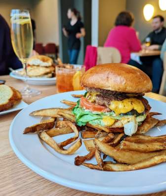A large burger with cheese, tomato, lettuce and sauce served on a white plate with fries. An old fashioned and glass of champagne are also on the table.