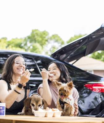 Two women and their dogs eat ice cream in front of a car.