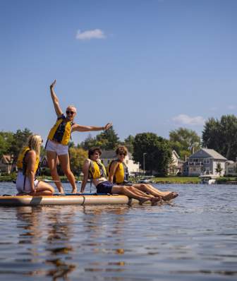 A group of four young adults sit on a paddleboard on the lake while one woman stands on the board with her arms in the air.