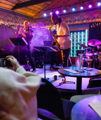 A man playing the saxophone along with other musicians perform on a stage in a dimly lit jazz club as the audience enjoys cocktails.