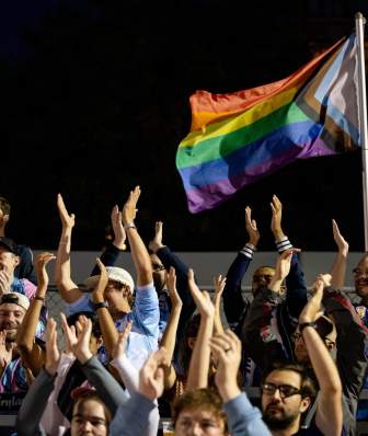 Pride flag waves above fans with arms raised at a Forward Madison FC soccer game