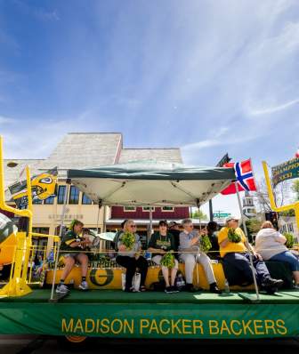 A green and yellow float that says Madison Packers Backers with several people sitting on it rides down the street during the Syttende Mai parade
