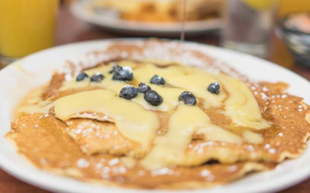 A close-up of a plate of pancakes topped with blueberries and syrup, with a blurred figure in the background pouring syrup. Glasses of orange juice are visible.