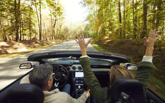 Couple driving convertible down nature filled road with roof down, and passenger has hands up