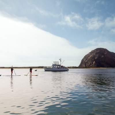 People paddle boarding in front of a fishing boat on the bay near Morro Rock