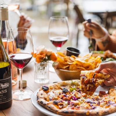 Table with wine bottles, glasses, and food. Person picking up a slice of pizza.